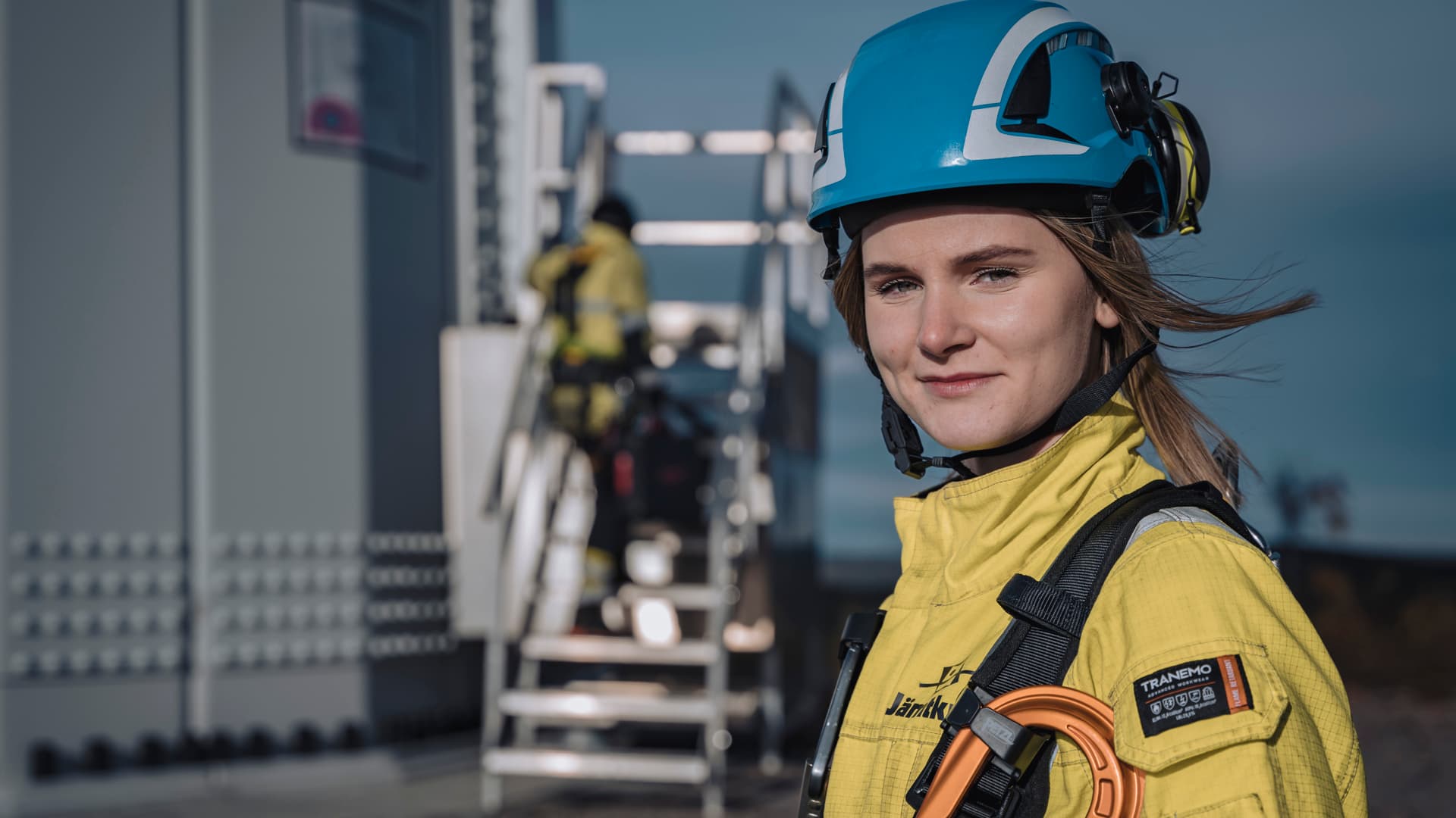 Porträtt av en kvinna i arbetskläder och skyddshjälm som står framför ett vindkraftverk.